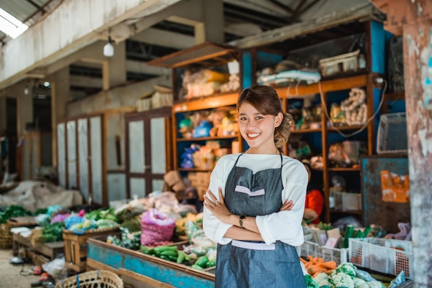 Asian female vegetable seller standing in front of vegetables stall smiling with crossed arm