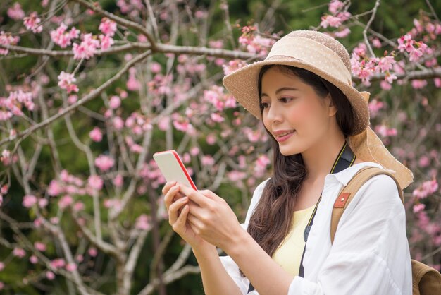 Asian female young lady standing under the tree and taking the photo with her cellphone.