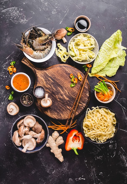 Asian food cooking concept. Empty wooden board, noodles, vegetables stir fry, shrimps, sauces, chopsticks.