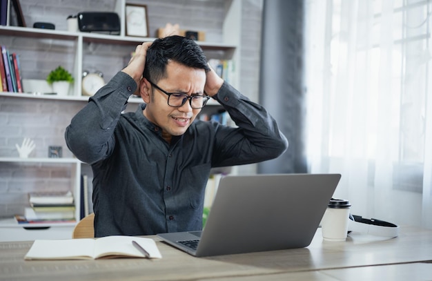 Asian freelance man wear glasses and hand over head having stressful depression sad time while working on laptop at home Depression man sad serios working from home Working at anywhere concept