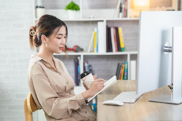 Asian freelance woman smiling holding cup of hot coffee and working on laptop computer on wooden table at home Entrepreneur woman working for her business at home Business work at home concept