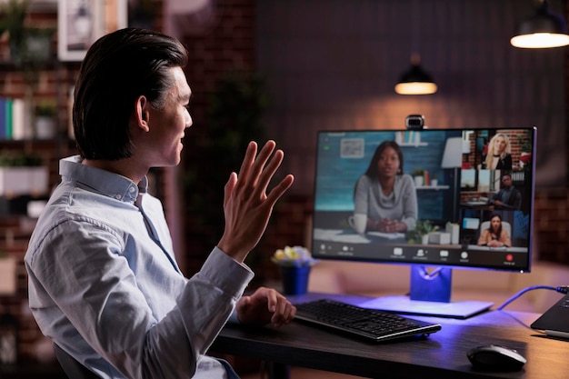 Asian freelancer waving at videocall conference with webcam, attending online teleconference meeting on computer. Talking to person on remote videoconference call, having telework conversation.