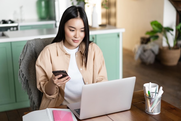 Asian girl applying for webinar girl with laptop and smartphone sitting at home working remote femal...