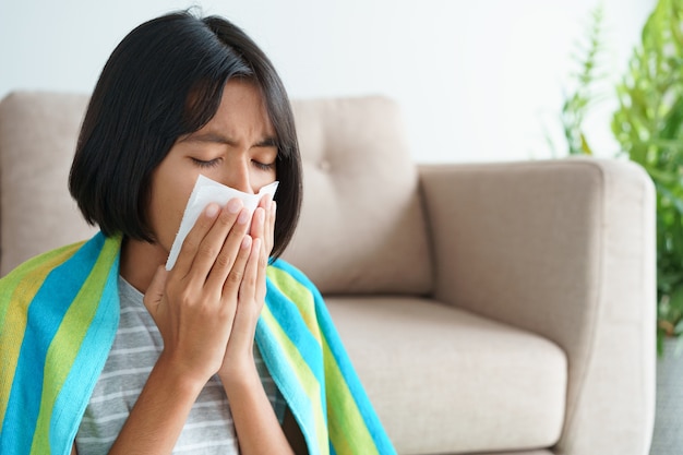 Asian girl having flu season and sneeze using paper tissues while sitting in the living room at home.