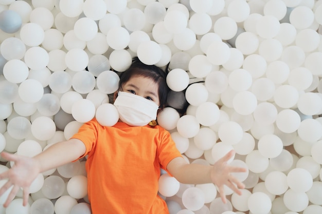 Asian girl play with white ball and wearing surgical mask on school or kindergarten yard or playground Little asian girl playing colorful ball at playground Child playing on outdoor playground