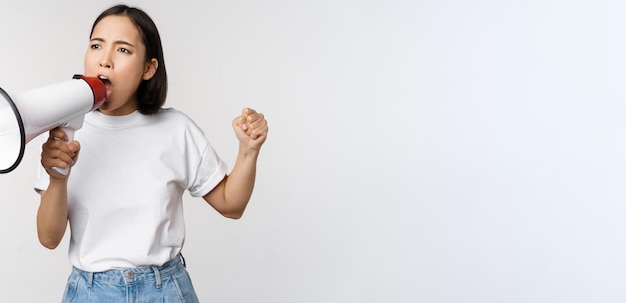 Asian girl shouting at megaphone young activist protesting using loud speakerphone standing over white background