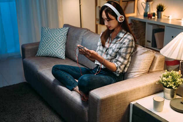 asian girl sitting on couch in casual wear is holding and watching online videos on her smartphone with headphone during nighttime in a cozy living room at home