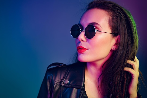 Asian girl in sunglasses and black leather jacket posing against dark blue background