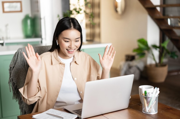 Asian girl on video conference saying hello waving hand at laptop camera working remote from home yo...