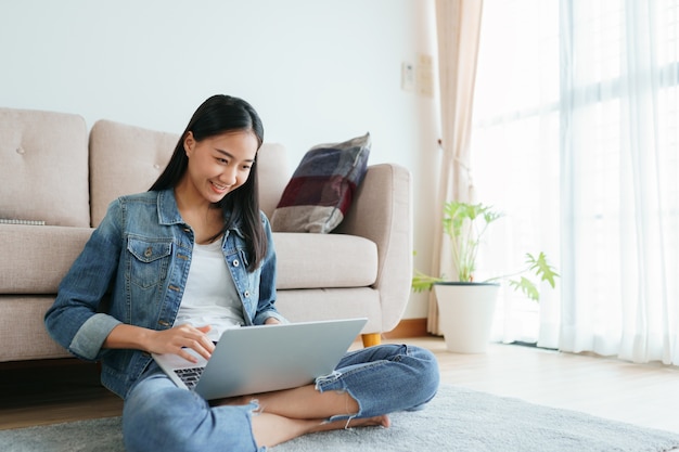 Asian girl wearing jeans using a laptop while sitting on the floor at home.