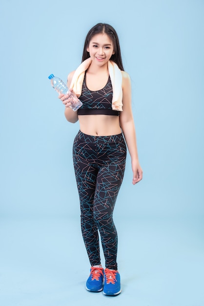 An Asian girl wearing a leggings and holding a water bottle to exercise on a pastel blue studio scene.