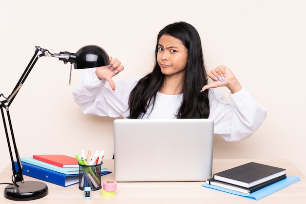 Asian girl in a workplace with a laptop on beige wall showing thumb down