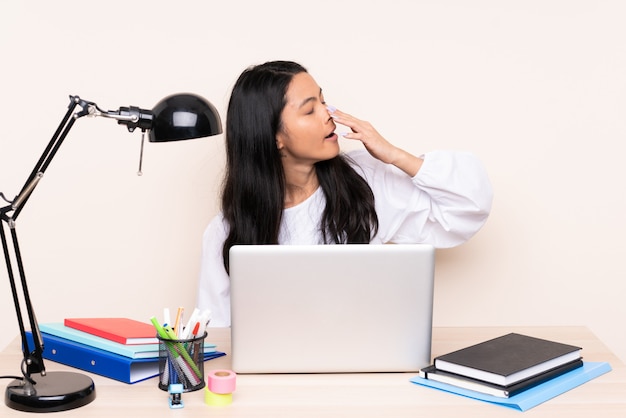 Asian girl in a workplace with a laptop on beige wall yawning and covering wide open mouth with hand