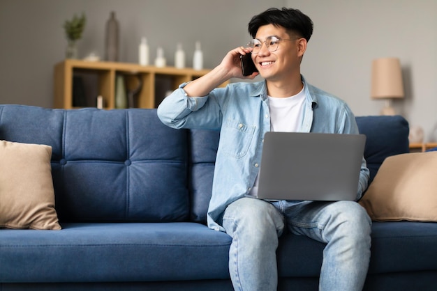 Asian guy in glasses with laptop communicating on phone indoor