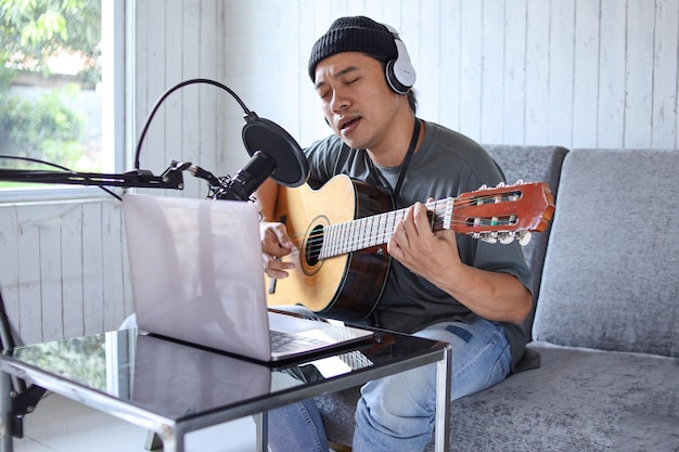 Asian guy playing the guitar recording audio podcast in studio using microphone and laptop. People a