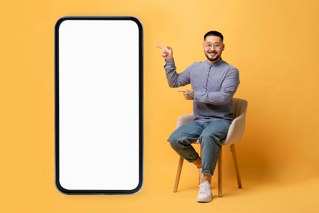 Asian guy pointing at smartphone with white screen while sitting on chair