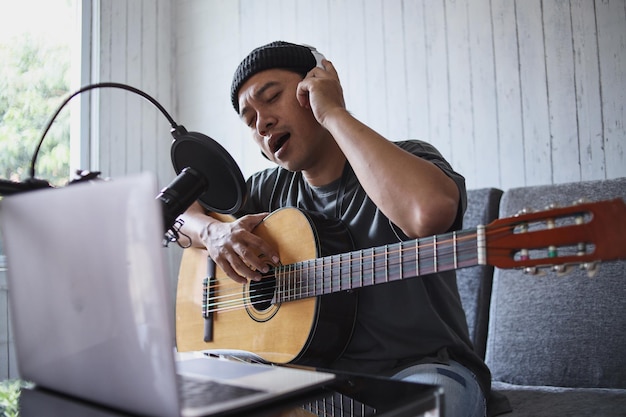 Asian guy singing and playing the guitar recording audio podcast in studio using microphone and lapt