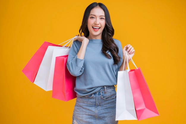 Asian happy female woman girl holds colourful shopping packages standing on yellow background studio shot Close up Portrait young beautiful attractive girl smiling looking at camera with bags