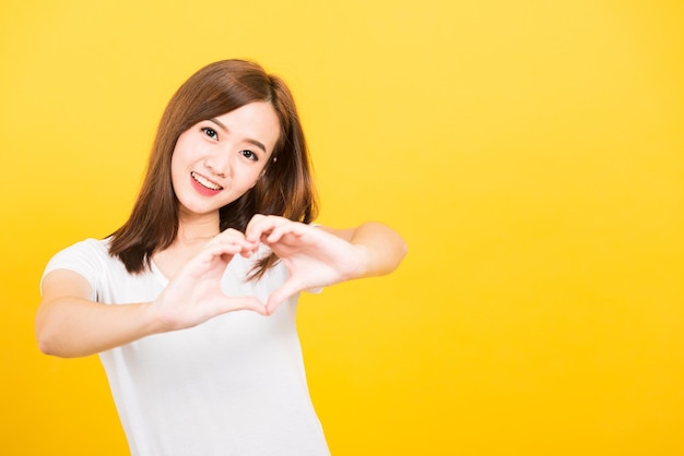 Asian happy portrait beautiful cute young woman teen smile standing make finger heart figure symbol shape sign with two hands looking to camera isolated, studio shot yellow background with copy space