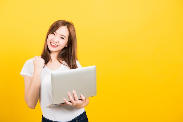 Asian happy portrait beautiful cute young woman teen smile standing wear t-shirt hold laptop computer and excited celebrating success looking to camera, studio shot yellow background with copy space