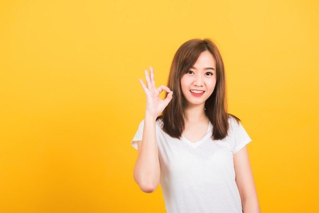 Asian happy portrait beautiful cute young woman teen standing wear t-shirt showing gesturing ok sign with fingers looking to camera isolated, studio shot on yellow background with copy space for text