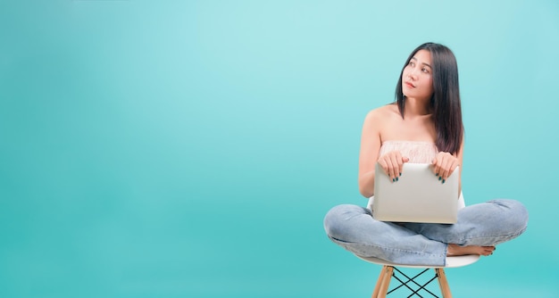 Asian happy portrait beautiful young woman sitting on chair smile her  holding laptop computer and looking to side on blue background with copy space for text