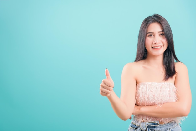 Asian happy portrait beautiful young woman standing her showing finger thumbs up and looking to camera isolated on blue background with copy space for text