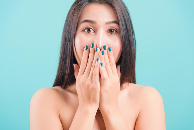 Asian happy portrait beautiful young woman standing smiling surprised excited her covering mouth with hands and looking to camera on blue background with copy space for text