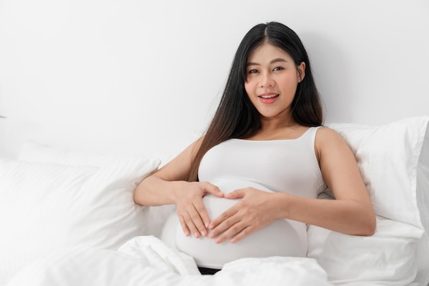 Asian happy pregnant woman is sitting on bed and and touching her belly. pregnancy, motherhood, people and expectation concept