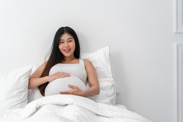 Asian happy pregnant woman is sitting on bed and and touching her belly pregnancy motherhood people and expectation concept