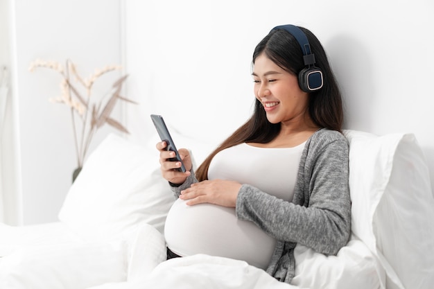 Asian happy pregnant woman is sitting on bed and and touching her belly and wear a headphone to listen the music. pregnancy, motherhood, people and expectation concept