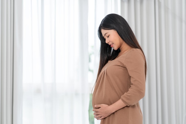 Asian happy pregnant woman is standing beside windows and touching her belly pregnancy motherhood people and expectation concept