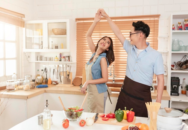 Asian husband and wife enjoy cooking salad and dancing together in kitchen room at home