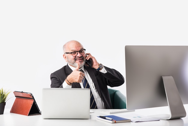 Asian Indian senior financial businessman sitting at his workstation or desk in front of a computer, laptop and tablet. Speaking on phone while doing some paperwork