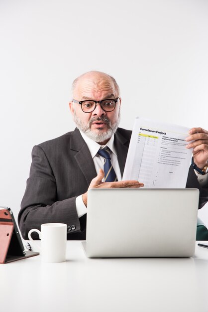 Asian Indian senior financial businessman sitting at his workstation or desk in front of a computer, laptop and tablet. Speaking on phone while doing some paperwork