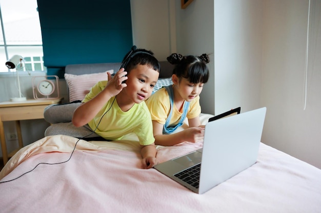 Asian kid boy using laptop and headphone for online study with video call teacher and have cute sister help him at bedroom