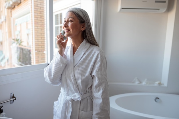 Asian lady doing face clean standing near the window in bathroom