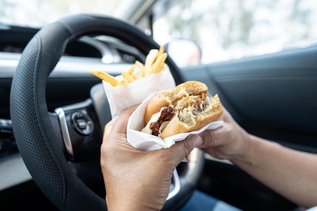 Asian lady holding hamburger and French fries to eat in car dangerous and risk an accident