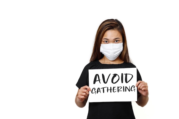 Asian lady standing with avoid gathering text front view in white plain wall Young lady looking straight wearing face mask for health and safety to avoid coronavirus