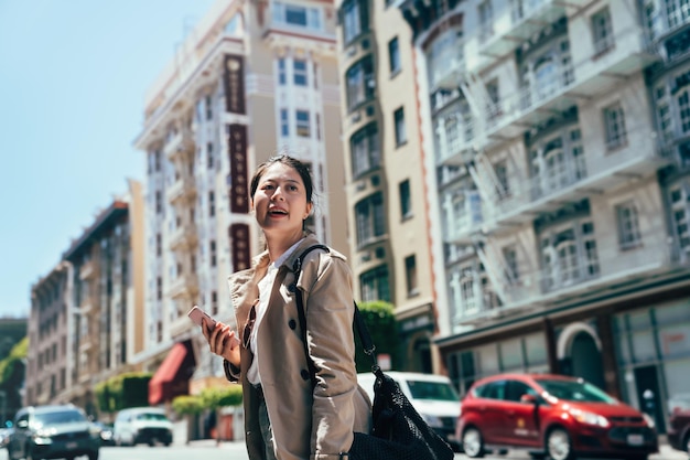 asian lady traveler holding cellphone carry backpack looking around searching tourist attraction standing on street on sunny day in san francisco. cars driving on road in background. tour in america