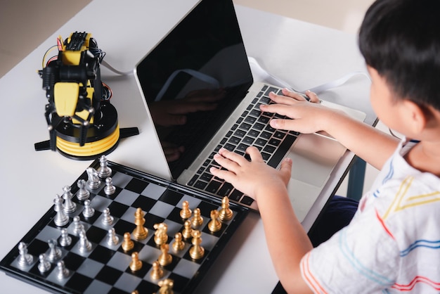 Asian little boy programming code to robot machine arm on laptop for play chess