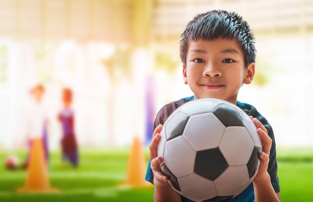 Asian little football boy with smile is holding a soccer ball with training ground backgorund.