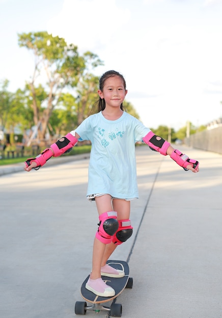 Asian little girl child skateboarder wearing safety and protective equipment playing on skateboard. Kid skateboarding on the road outdoors