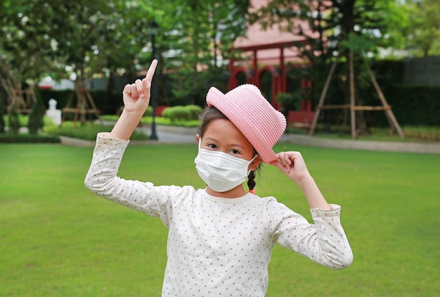 Asian little girl child wearing medical mask and straw hat with pointing one forefinger up due covid19 on the garden