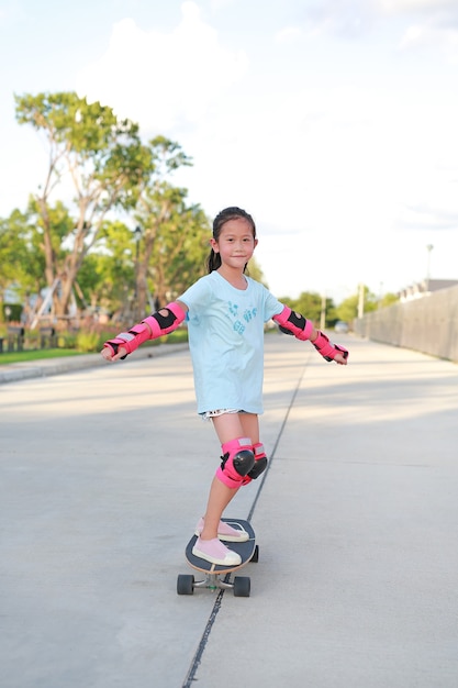 Asian little girl child wearing safety and protective equipment skating on a skateboard. Kid riding on skateboard outdoors at the street