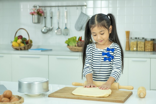 Asian little girl cook rolls the dough for pizza with a wooden rolling pin on a black table sprinkled with flour.
