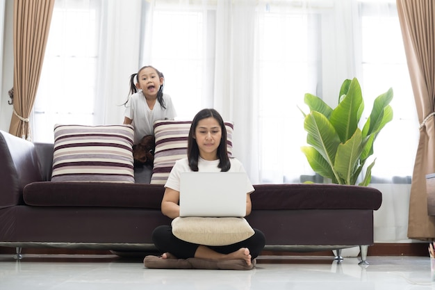 Asian little girl looking how mother working with laptop in living room