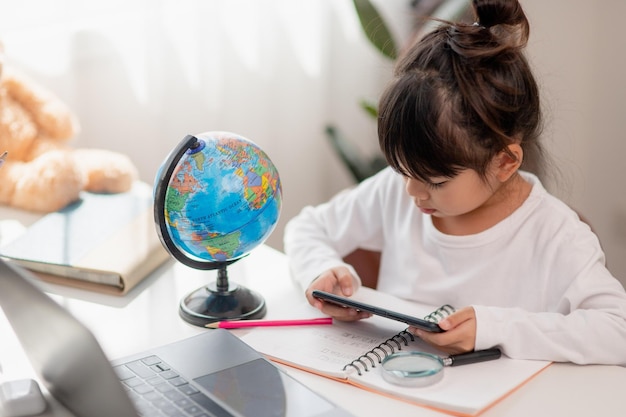 Asian little girl using a phone close up distracted from studying sitting at a table with notebooks a pretty child having fun with a smartphone watching the webinar homeschooling