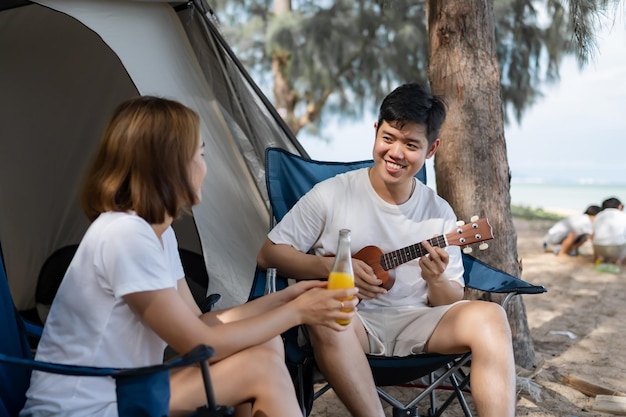 Asian love couple camping on the summer beach. Man playing Ukulele and together with woman sitting outside the tent.