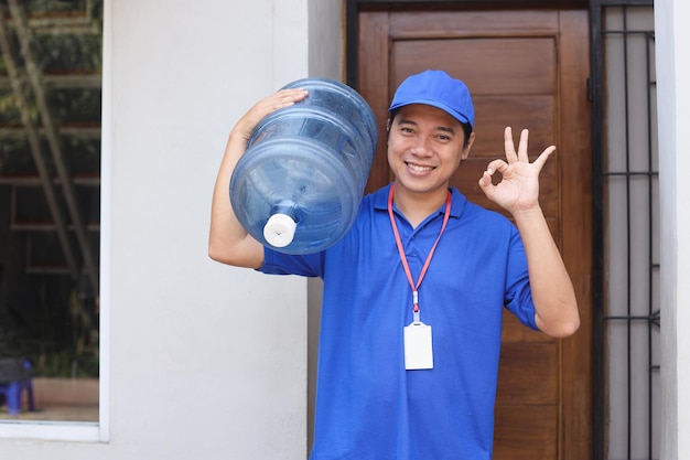 Asian male courier carry bottle with clean water on shoulder while showing okay sign